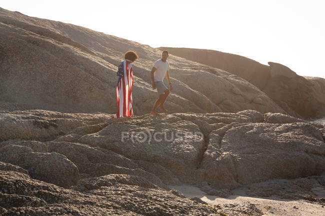 Vorderansicht eines afrikanisch-amerikanischen Paares, das zusammen auf einem Felsen mit amerikanischer Flagge in der Nähe des Meeres genießt — Stockfoto