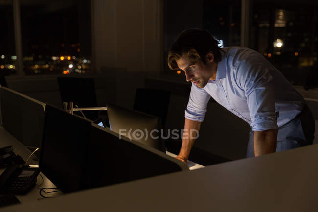 Vista lateral del apuesto joven ejecutivo de pie y trabajando en el escritorio en una oficina moderna. Está trabajando hasta tarde. - foto de stock