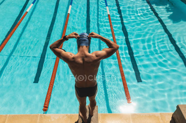 Высокий угол обзора кавказского пловца, стоящего на стартовом блоке и одетого в плавательные очки у бассейна на солнце — стоковое фото