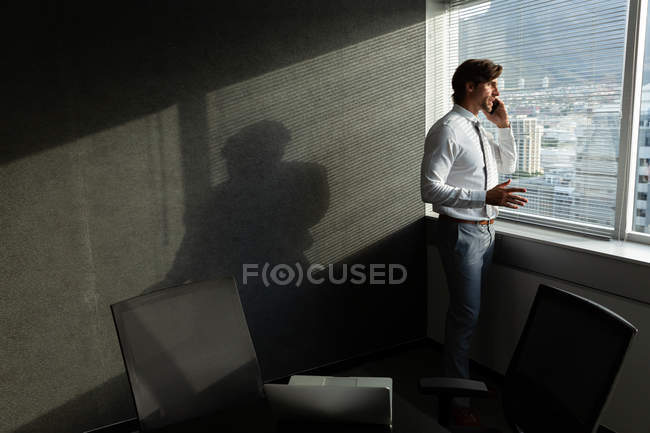 Vista lateral de guapo joven ejecutivo hablando por teléfono móvil mientras está de pie cerca de la ventana en una oficina moderna - foto de stock