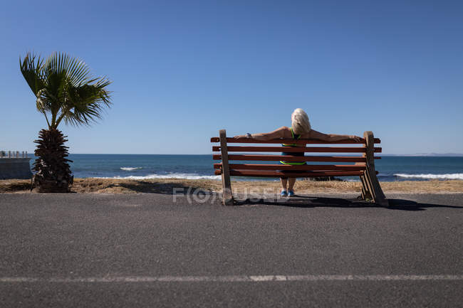 Вид сзади активной пожилой женщины, отдыхающей на скамейке рядом с пляжем под солнцем — стоковое фото