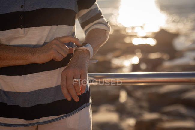 Vista de cerca de una mujer mayor activa usando su smartwatch mientras se apoya contra un carril de guardia en un paseo marítimo en la noche - foto de stock