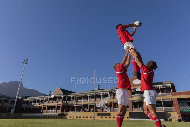 Низький кут зору чоловіків-регбістів, які ловили м'яч у повітрі під час дотику на стадіоні в сонячний день — стокове фото
