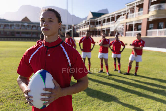 Vue de face d'un joueur de rugby tenant la balle de rugby et regardant la caméra dans le terrain de rugby par une journée ensoleillée — Photo de stock