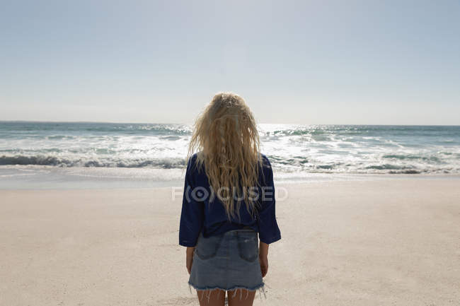 Vista trasera de la hermosa mujer rubia de pie en la playa en un día soleado. Ella está mirando al océano - foto de stock