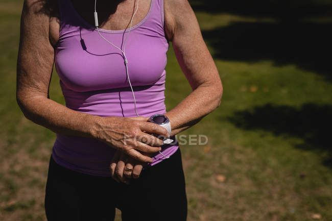 Середина активної старшої жінки, використовуючи смарт-годинник, слухаючи музику з навушниками в парку в сонячний день — стокове фото