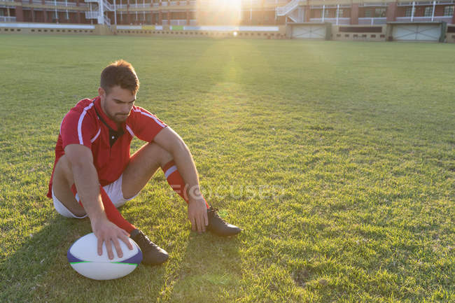 Vista frontal de un hombre reflexivo jugador de rugby caucásico sentado con pelota de rugby en el suelo - foto de stock