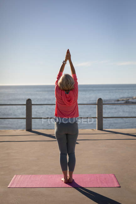 Rückansicht einer aktiven Seniorin, die Yoga auf einer Fitnesskarte auf einer Promenade am Meer macht — Stockfoto