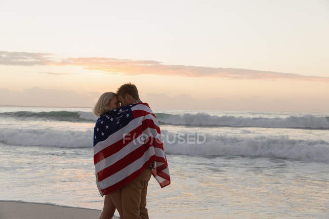 Vista laterale della giovane coppia romantica avvolta nella bandiera americana in spiaggia. Si abbracciano. — Foto stock