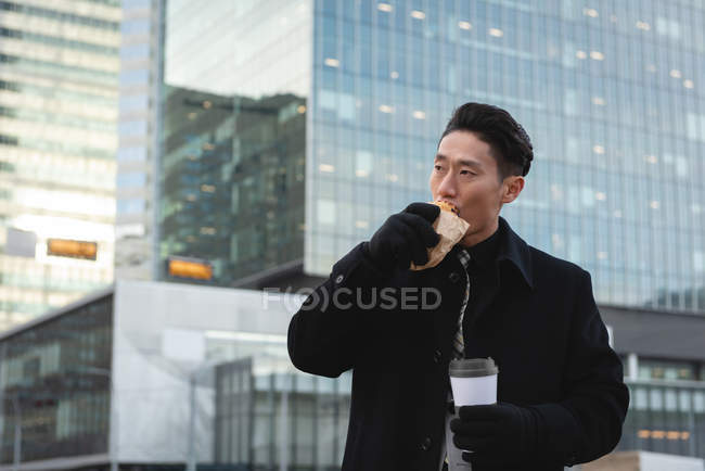 Vista frontal de un joven empresario comiendo comida y sosteniendo café mientras está parado en la calle de la ciudad - foto de stock