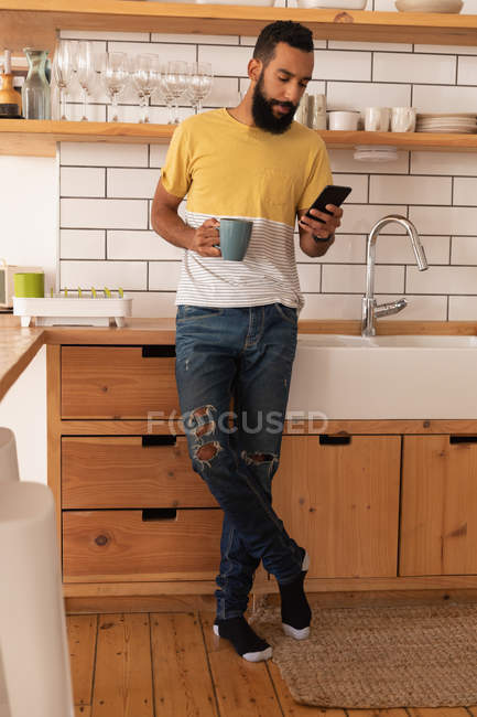 Передній вигляд змішаної раси людини, що використовує мобільний телефон, маючи чашку кави вдома в кухонній кімнаті — стокове фото