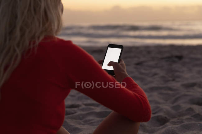 Vue arrière de la femme blonde utilisant un téléphone portable à la plage. Elle est assise sur le sable — Photo de stock