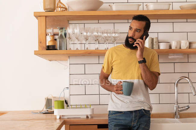 Вид спереду змішаної раси Чоловік розмовляє по мобільному телефону, стоячи вдома в кухонній кімнаті — стокове фото