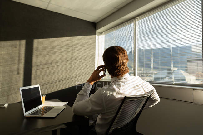 Visão traseira do jovem executivo sentado à mesa e falando no telefone celular em um escritório moderno — Fotografia de Stock