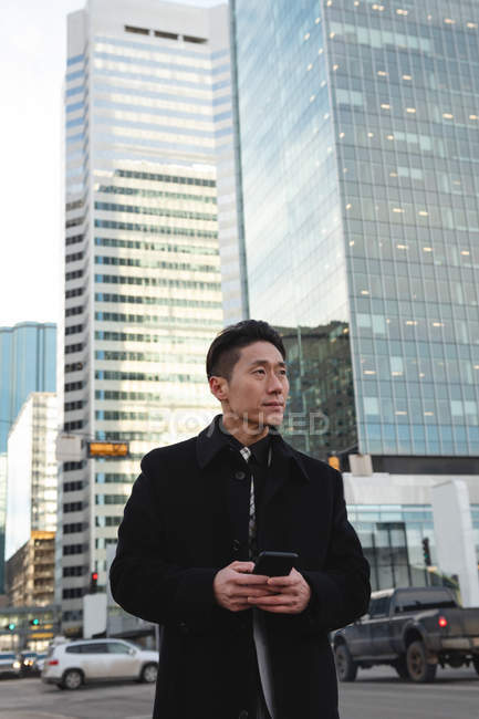 Вид спереду вдумливого молодого азіатського бізнесмена, який використовує мобільний телефон, стоячи на вулиці з будівлею позаду — стокове фото
