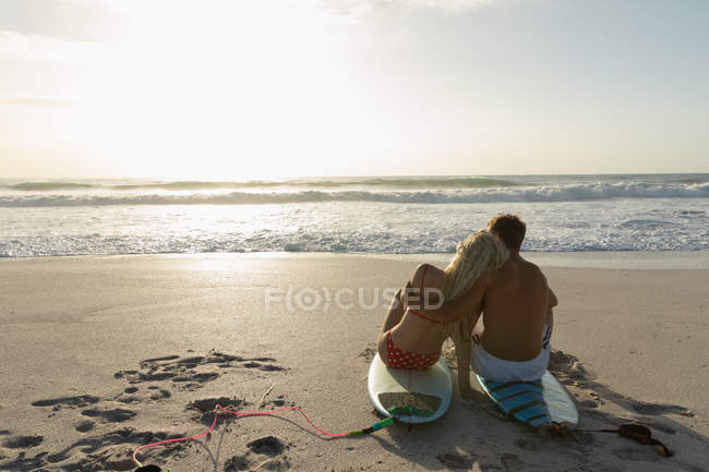 Rückansicht eines glücklichen Paares, das sich an einem sonnigen Tag auf einem Surfbrett am Strand entspannt. Sie umarmen sich — Stockfoto