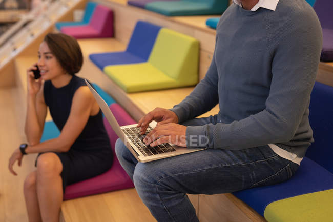 Seitenansicht eines Geschäftsmannes, der seinen Laptop benutzt, während eine Geschäftsfrau im Büro auf ihrem Mobiltelefon spricht — Stockfoto