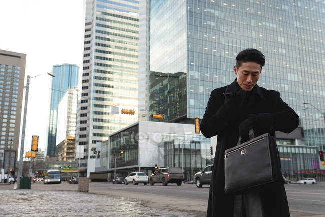 Vista de bajo ángulo del joven empresario asiático mirando smartwatch mientras habla por teléfono móvil en la calle en la ciudad - foto de stock