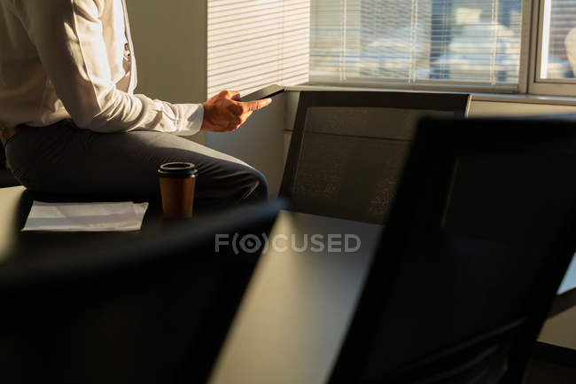 Середина чоловічої виконавчої влади, що працює на мобільному телефоні, сидячи на столі і насолоджуючись чашкою кави в сучасному офісі — стокове фото