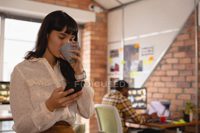 Вид спереду бізнес-леді, використовуючи мобільний телефон, маючи чашку кави в офісі — стокове фото