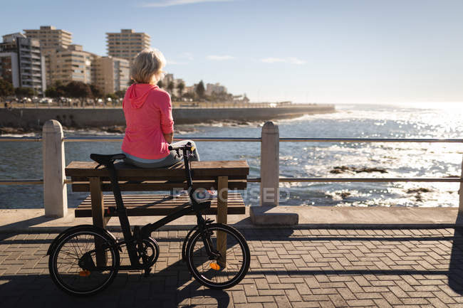 Rückansicht einer aktiven Seniorin, die es sich auf einer Bank gemütlich macht und auf der Promenade in der Sonne auf das Meer blickt — Stockfoto