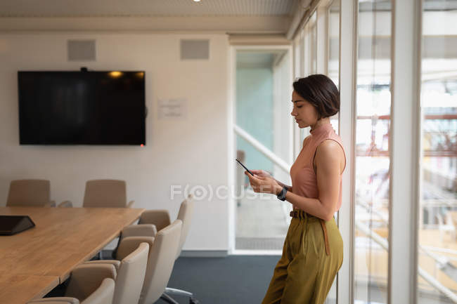 Вид сбоку на азиатскую деловую женщину с мобильного телефона, стоящую у окна в офисе — стоковое фото