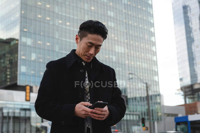 Vue de face du jeune homme d'affaires asiatique utilisant un téléphone portable dans la rue de la ville avec un bâtiment derrière lui — Photo de stock