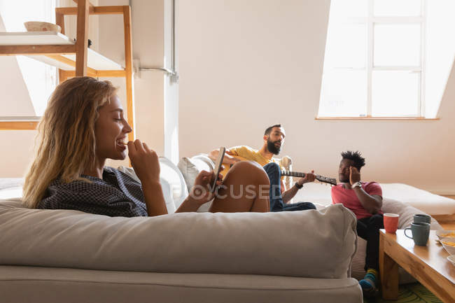 Вид сбоку на женщину, использующую мобильный телефон, в то время как мужчина играет на гитаре в гостиной дома — стоковое фото