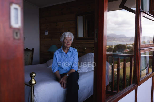 Vista frontal de una mujer mayor activa pensativa sentada en la cama y mirando a través de la ventana en el dormitorio en casa - foto de stock