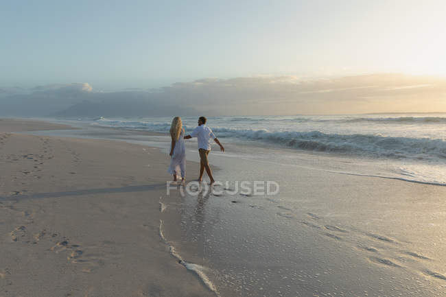 Vista trasera de la joven pareja de amor cogida de la mano mientras camina en la playa en un día soleado - foto de stock