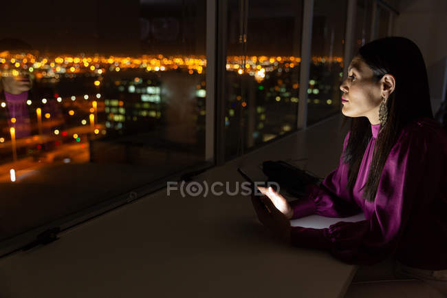 Seitenansicht der schönen jungen weiblichen Führungskraft gemischter Rassen mit digitalem Tablet, die durch ein Fenster in einem modernen Büro blickt — Stockfoto