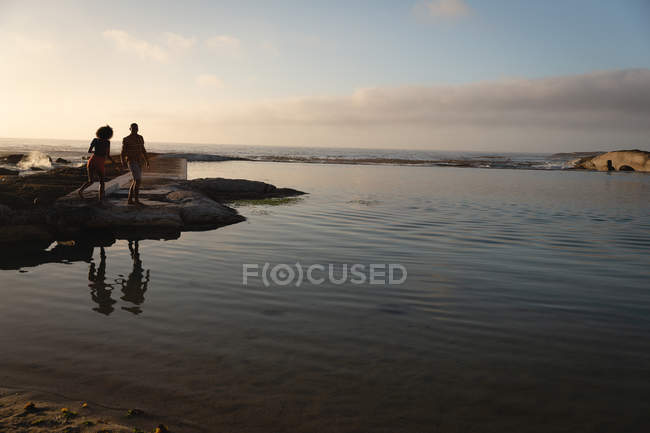 Vista a distanza della coppia afro-americana in piedi e rilassante vicino al mare al tramonto. Sono in piedi sulla roccia — Foto stock