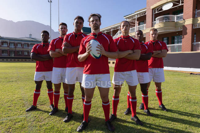Vista frontale di una squadra di giocatori di rugby multietnico maschile in piedi con una palla da rugby nel terreno di rugby in una giornata di sole — Foto stock