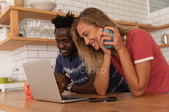 Vista laterale della coppia multietnica in piedi e al lavoro su laptop in cucina a casa — Foto stock