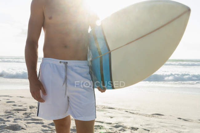 Средняя часть молодого серфера с доской для серфинга, стоящей на пляже в солнечный день — стоковое фото