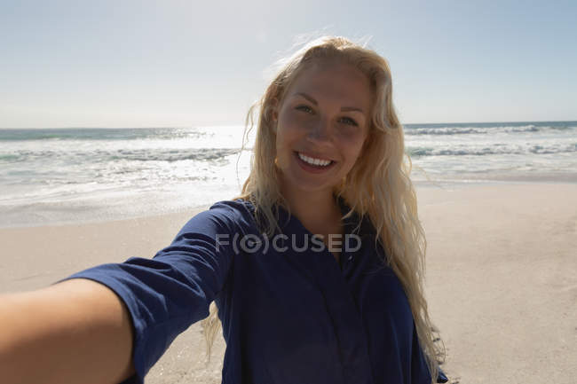 Vorderansicht der schönen blonden Frau, die an einem sonnigen Tag am Strand steht. Sie macht ein Selfie — Stockfoto