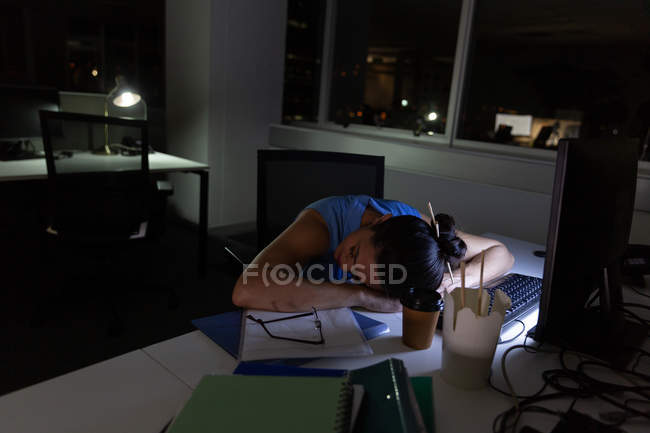 Vista frontal de una joven ejecutiva de raza mixta durmiendo en un escritorio en una oficina moderna - foto de stock