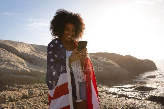 Vista frontal da mulher afro-americana de pé e usando telefone celular com bandeira americana perto do lado do mar — Fotografia de Stock