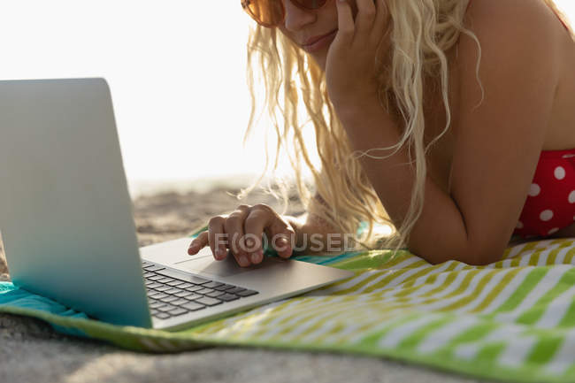 Sección media de la mujer rubia usando el ordenador portátil mientras está acostado en la playa en un día soleado - foto de stock