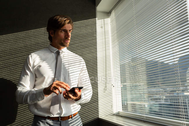 Vista frontal del apuesto joven ejecutivo masculino con teléfono móvil mirando por la ventana en una oficina moderna - foto de stock