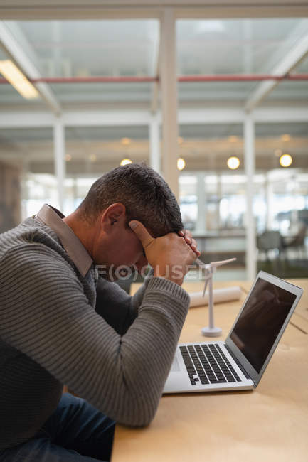 Вид сбоку на печального бизнесмена, сидящего за столом с ноутбуком и держащего голову руками — стоковое фото