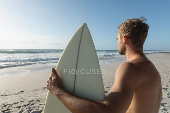 Vista lateral del joven surfista masculino con una tabla de surf de pie en una playa en un día soleado. Él está observando las olas - foto de stock