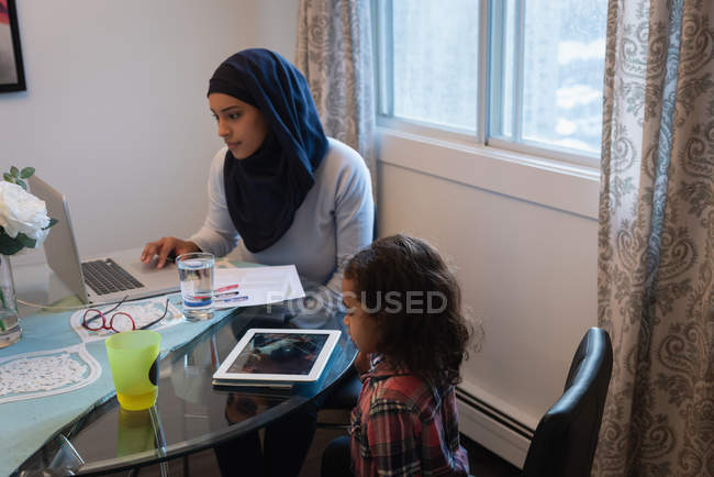 Visão lateral da mãe de raça mista usando hijab usando laptop enquanto filha olhando para tablet digital em casa. Eles estão sentados em torno de uma mesa na sala de estar — Fotografia de Stock