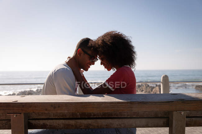 Seitenansicht eines afrikanisch-amerikanischen Paares in romantischer Stimmung, das auf einer Holzplanke in Meeresnähe sitzt — Stockfoto