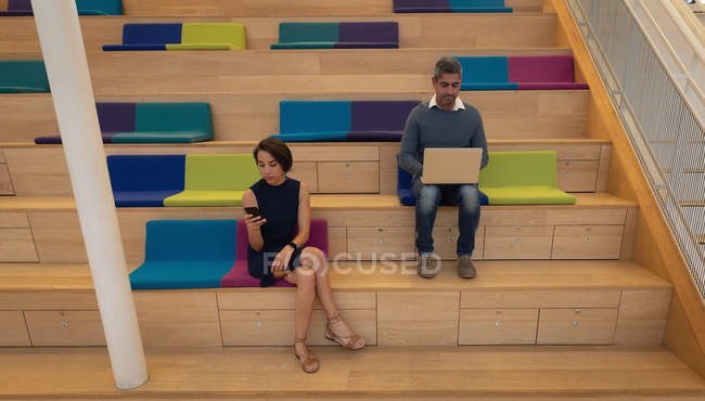Front view of a Caucasian businessman using a laptop while a young businesswoman use her mobile phone and sit on stairs — Stock Photo