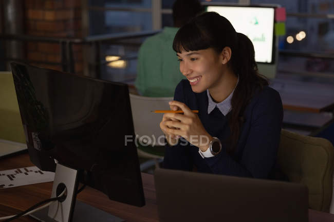 Vue de face de femme d'affaires asiatique souriant tout en regardant l'ordinateur dans le bureau — Photo de stock