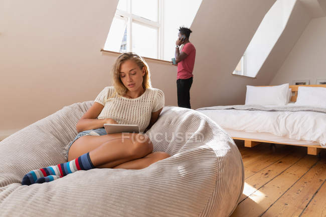 Vista frontal de la mujer utilizando la tableta digital en el sofá, mientras que el hombre habla en el teléfono móvil en casa - foto de stock