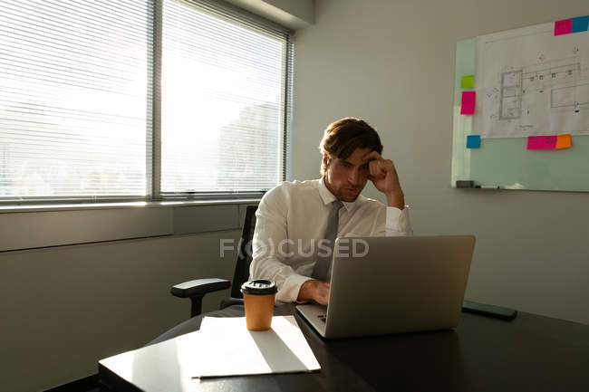 Вид спереду красивого молодого керівника чоловічої статі сидить за столом і працює на ноутбуці в сучасному офісі — стокове фото