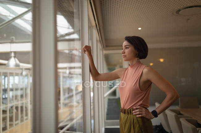 Vista lateral de uma empresária asiática pensativa de pé no escritório e olhando pela janela — Fotografia de Stock