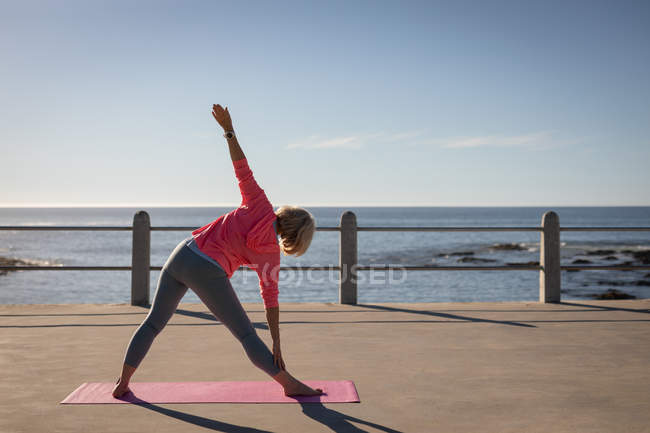Vista trasera de una mujer mayor activa practicando ejercicios de fitness en una alfombra de fitness en un paseo marítimo bajo el sol frente a la playa - foto de stock